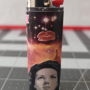May include: A black and silver lighter with a colorful design featuring a woman's face, a galaxy, and red lips. The lighter has the word "BIC" printed on it.