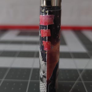May include: A silver lighter with a black and red design. The design features a collage of images, including a person's face, a galaxy, and a pink and red stripe.