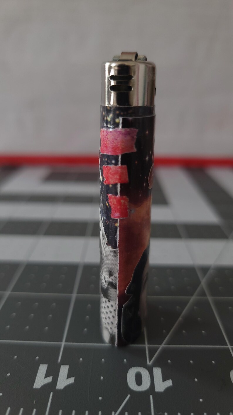 May include: A silver lighter with a black and red design. The design features a collage of images, including a person's face, a galaxy, and a pink and red stripe.