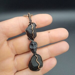Puede incluir: Un colgante de obsidiana negra con un dije de calavera, envuelto en alambre de cobre. El colgante cuelga de una cadena de cobre.