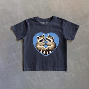Puede incluir: Una camiseta gris oscuro con un dise&ntilde;o de coraz&oacute;n azul que presenta dos mapaches en su interior. Los mapaches se miran y parecen estar abraz&aacute;ndose.