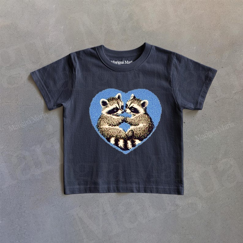 Puede incluir: Una camiseta gris oscuro con un dise&ntilde;o de coraz&oacute;n azul que presenta dos mapaches en su interior. Los mapaches se miran y parecen estar abraz&aacute;ndose.