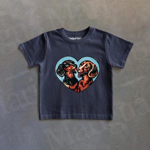 Puede incluir: Camiseta azul marino con un dise&ntilde;o en forma de coraz&oacute;n con dos perros salchicha bes&aacute;ndose, sobre un fondo azul claro. El texto "Marigua Maria" est&aacute; impreso en la camiseta.