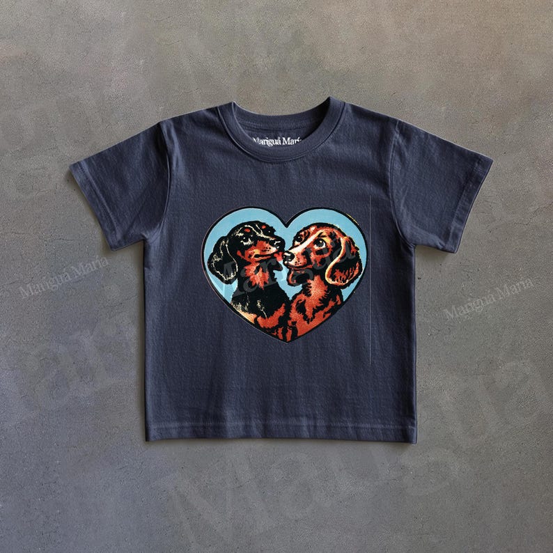 Puede incluir: Camiseta azul marino con un dise&ntilde;o en forma de coraz&oacute;n con dos perros salchicha bes&aacute;ndose, sobre un fondo azul claro. El texto "Marigua Maria" est&aacute; impreso en la camiseta.