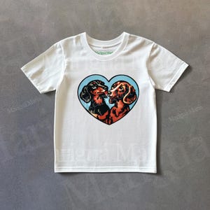 Puede incluir: Camiseta blanca con un dise&ntilde;o en forma de coraz&oacute;n de dos perros salchicha. Uno es negro y marr&oacute;n, el otro es marr&oacute;n. Est&aacute;n nariz con nariz sobre un fondo azul claro. La camiseta tiene mangas cortas.