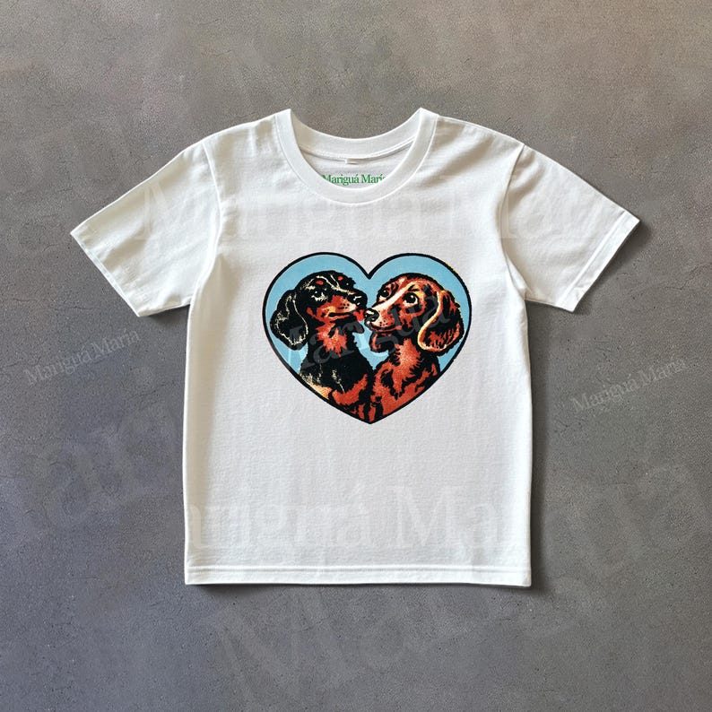 Puede incluir: Camiseta blanca con un dise&ntilde;o en forma de coraz&oacute;n de dos perros salchicha. Uno es negro y marr&oacute;n, el otro es marr&oacute;n. Est&aacute;n nariz con nariz sobre un fondo azul claro. La camiseta tiene mangas cortas.