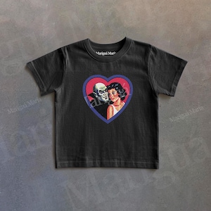 Puede incluir: Camiseta negra con un gráfico en forma de corazón que representa una ilustración vintage de un hombre calvo y una mujer. El gráfico es rojo, azul y blanco. El texto "Mariguá Maria" está impreso en la camiseta.