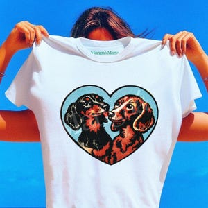 Puede incluir: Camiseta blanca con un dise&ntilde;o en forma de coraz&oacute;n que muestra a dos perros salchicha bes&aacute;ndose. Los perros son de color negro y marr&oacute;n, sobre un fondo azul claro. El texto "Marigu&aacute; Maria" est&aacute; impreso en el cuello.