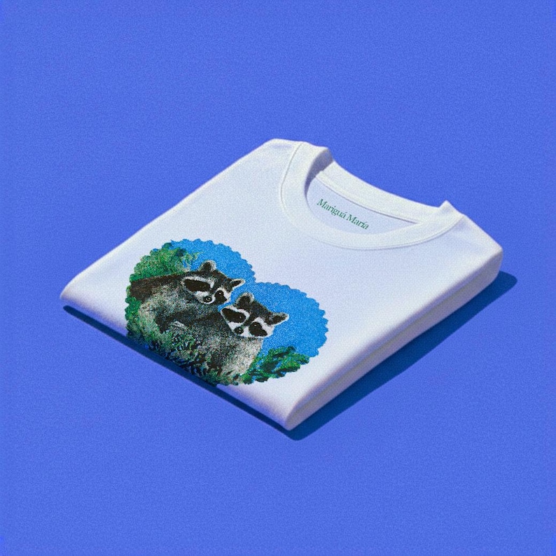May include: White t-shirt with a graphic of two raccoons inside a blue heart-shaped design. The shirt has the text "Marigua Maria" printed on the collar. The shirt is folded on a blue surface.