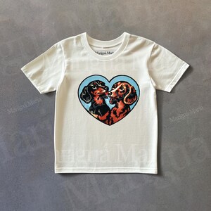 Puede incluir: Camiseta blanca con un dise&ntilde;o en forma de coraz&oacute;n con dos perros salchicha. Los perros son marrones y negros, sobre un fondo azul claro. El texto "Marigu&aacute; Maria" est&aacute; impreso en la camiseta.