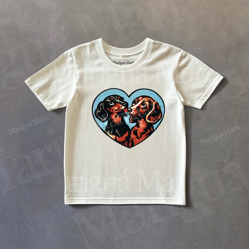 Puede incluir: Camiseta blanca con un dise&ntilde;o en forma de coraz&oacute;n con dos perros salchicha. Los perros son marrones y negros, sobre un fondo azul claro. El texto "Marigu&aacute; Maria" est&aacute; impreso en la camiseta.