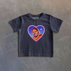 May include: Dark gray t-shirt featuring a heart-shaped graphic with a Chewbacca illustration. The graphic is outlined in pink and filled with a blue background. The text "Mariqua Maria" is printed on the shirt.