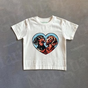 Puede incluir: Camiseta de color crema con un dise&ntilde;o en forma de coraz&oacute;n con dos perros salchicha sobre un fondo azul. Los perros salchicha est&aacute;n representados en negro y marr&oacute;n, con sus narices toc&aacute;ndose. La camiseta tiene mangas cortas y cuello redondo.