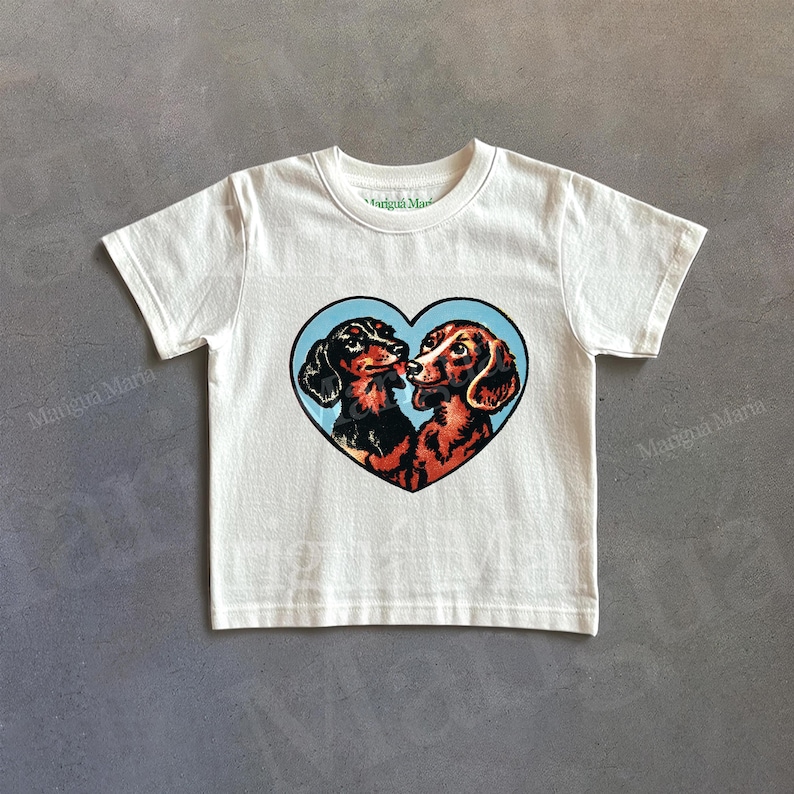Puede incluir: Camiseta de color crema con un dise&ntilde;o en forma de coraz&oacute;n con dos perros salchicha sobre un fondo azul. Los perros salchicha est&aacute;n representados en negro y marr&oacute;n, con sus narices toc&aacute;ndose. La camiseta tiene mangas cortas y cuello redondo.