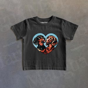 Puede incluir: Una camiseta gris oscuro con un dise&ntilde;o en forma de coraz&oacute;n que muestra a dos perros salchicha bes&aacute;ndose. Los perros est&aacute;n sobre un fondo azul claro. La camiseta tiene cuello redondo y mangas cortas.