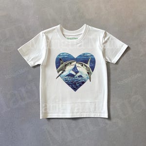 May include: White t-shirt featuring a graphic of two sharks kissing within a blue heart. The text "Margaux Maris" is printed on the shirt.