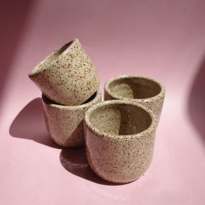 May include: A set of three small ceramic cups with a speckled beige finish. Two cups are positioned upright on the surface, while one cup is tilted and resting on top of another, showcasing the smooth, rounded edges and the consistent brown speckle pattern.