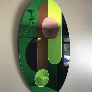 May include: A green, brown, and yellow oval mirror with a black background. The mirror is divided into sections with different colored shapes. The mirror is hanging on a wall.