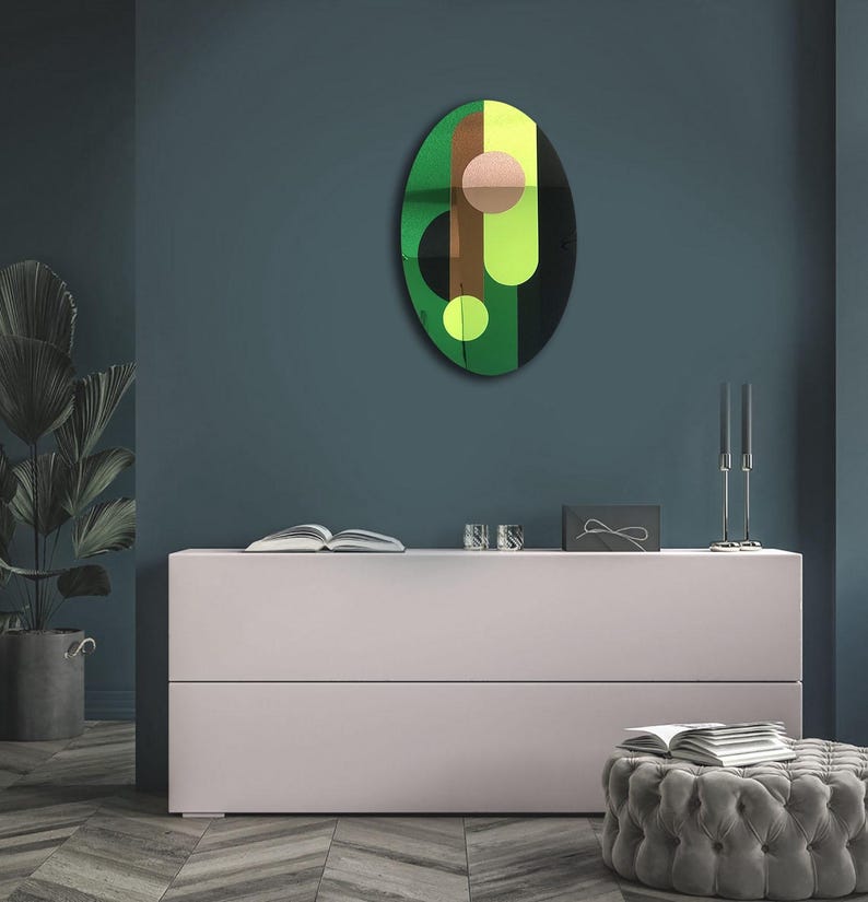 May include: A green, yellow, brown, and black abstract oval wall art piece with geometric shapes. The art piece is hanging on a dark blue wall above a white dresser.