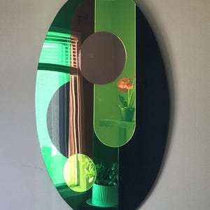 May include: An oval-shaped mirror with a green and black frame. The mirror is divided into sections with different shapes and sizes. The mirror reflects a window with blinds, a plant, and a flower.