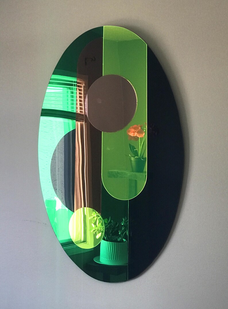 May include: An oval-shaped mirror with a green and black frame. The mirror is divided into sections with different shapes and sizes. The mirror reflects a window with blinds, a plant, and a flower.