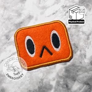 May include: Orange embroidered patch with a cartoon face design. The patch features black oval eyes, a black downward-pointing mouth, and a gold border. The patch is labeled "Physical Product".