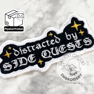 May include: Embroidered patch with the text "distracted by SIDE QUESTS" in white script on a black background, outlined in white. Gold star accents are included. A "Physical Product" sticker and a "Mochi Makes Embroidery" sticker are also visible.