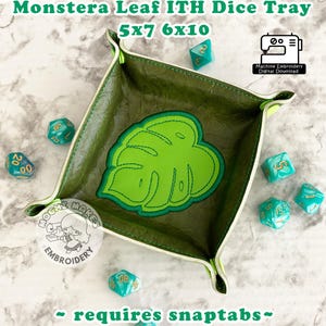May include: Green dice tray featuring a monstera leaf design. The tray is square with a stitched border and a green leaf in the center. The text "Monstera Leaf ITH Dice Tray" and "5x7 6x10" are visible. The text "requires snaptabs" is also visible.
