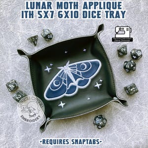 May include: A black leather dice tray with a blue and white embroidered lunar moth design. The tray is 5x7 inches and 6x10 inches. The tray is surrounded by black dice with white numbers. The text "LUNAR MOTH APPLIQUE ITH 5X7 6X10 DICE TRAY" is at the top of the image. The text "MOCHI MAKES EMBROIDERY" is at the bottom of the image. The text "REQUIRES SNAPTABS" is at the bottom of the image.