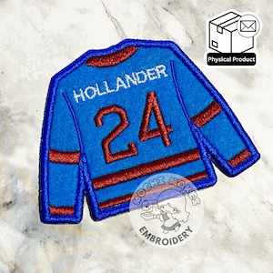Peut inclure: Écusson de maillot de hockey brodé bleu et rouge, avec le nom "HOLLANDER" et le numéro "24". L'écusson a une base bleue avec des détails rouges. Le texte est en blanc.