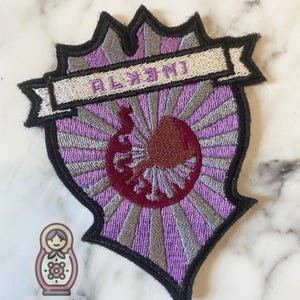 May include: Purple and silver embroidered patch with a stylized sunburst design and a red and silver crest. The patch has a white banner with the word "ALXMY" embroidered on it.