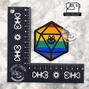 Può includere: Toppa ricamata a forma di D20 con sfumatura arcobaleno e faccia di gatto nero. La toppa è larga circa 7.6 cm. L'immagine include un righello nero con segni decorativi e il testo "Machine Embroidery Digital Download".