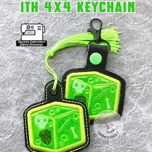 Könnte beinhalten: Grüner gallertartiger Würfel-Schlüsselanhänger mit einem schwarzen Lederband und einem grünen Quasten. Der Schlüsselanhänger ist ein Maschinenstickdesign, und das Bild enthält den Text "GELATINOUS CUBE ITH 4X4 KEYCHAIN" und "MOCHI MAKES EMBROIDERY".