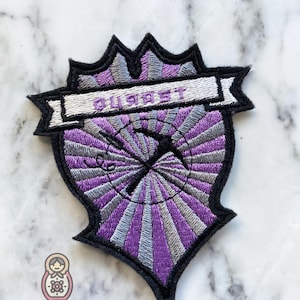 May include: Purple and silver embroidered patch with a sunburst design and the word "QUEST" in a banner at the top.