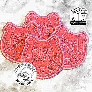 May include: Several pink horseshoe-shaped embroidered patches. Each patch has the text "GOOD LUCK BABE" and heart details. The patches are a physical product.