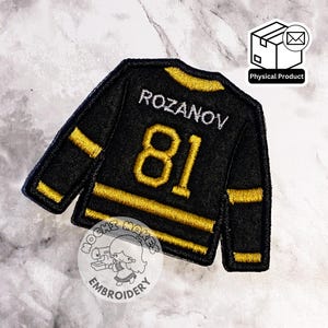 May include: Black and gold hockey jersey patch with the name "Rozanov" and the number "81" embroidered in gold. The patch has gold stripes on the sleeves and collar. The patch is a physical product.