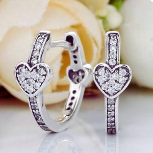 Puede incluir: Un par de pendientes de aro de plata con un dije en forma de corazón en la parte frontal de cada aro. Los corazones y la superficie frontal de los aros están incrustados con pequeñas piedras claras y brillantes en engaste pavé.