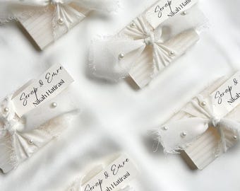 May include: Several small, white rectangular objects are adorned with white fabric bows and faux pearls. Each object has a small tag with the text "Serap & Emre Nikah Hatirasi" in black script. The items are arranged on a white, textured surface.