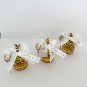 May include: Three small glass jars filled with honey, each tied with a white fabric ribbon and a small sprig of dried flowers. A wooden honey dipper is attached to each jar. The jars have a handwritten label that reads "Megan & Tim".