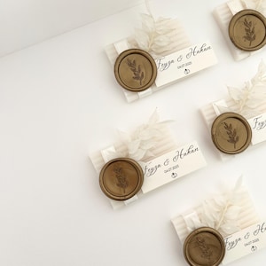 May include: Three white soap favours with gold wax seals and a white ribbon. Each soap favour has a small white tag with the text "Fevza & Hakan 04.07.2025" and a small gold ring icon.