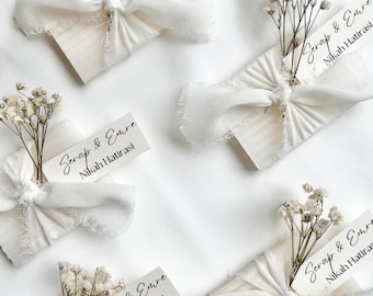 May include: Arrangement of white favors, each wrapped with a cream-colored ribbon and a sprig of dried flowers. Each favor has a small tag with the text "Serap & Emre Nikah Hatirasi" in black script. The favors are arranged on a white surface.
