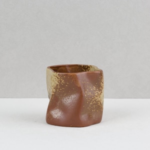 May include: A small, hand-formed ceramic cup with an irregular, dimpled shape. The exterior features a two-tone glaze, with a smooth, solid reddish-brown base transitioning into a textured, speckled beige finish toward the top rim.