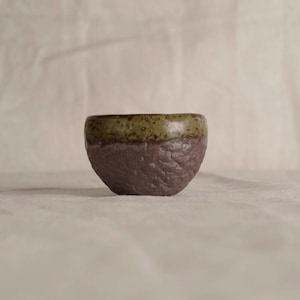 May include: A small, handcrafted ceramic bowl with a textured, dark purple exterior and a green-brown rim. The bowl has a rounded shape and is likely used for serving or display.