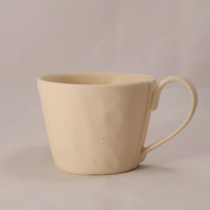 Könnte beinhalten: Cremefarbene Keramiktasse mit strukturierter Oberfläche und gebogenem Henkel. Die Tasse hat eine weite Öffnung und einen leicht konischen Körper. Der Griff ist an der Seite der Tasse befestigt.