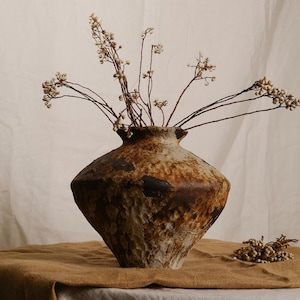 May include: A textured ceramic vase with a wide base and a narrow neck, filled with dried branches bearing small, white berries. The vase has a mottled brown and tan finish, and sits on a brown cloth. The vase is approximately 30 cm tall.