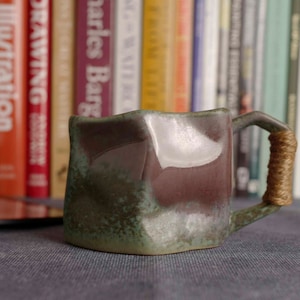 May include: A green and brown ceramic mug with a unique, uneven shape and a rope handle. The mug is sitting on a grey surface.