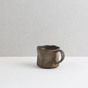 May include: A dark brown ceramic mug with a textured, uneven surface and a wavy rim. The mug features a metallic, mottled finish with subtle hints of bronze and black, and includes a curved handle attached to the side of the vessel.