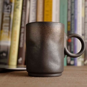 Könnte beinhalten: Eine dunkelbraune Keramiktasse mit rundem Henkel. Die Tasse hat eine strukturierte Oberfläche mit einem Goldverlauf am Rand. Die Tasse ist zylindrisch mit leicht ausgestelltem Boden. Bücher sind im Hintergrund.