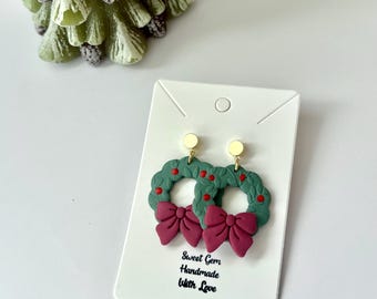 May include: A pair of festive earrings featuring a green wreath design with red berry accents and a burgundy bow. The earrings are attached to gold-colored studs. The card reads "Sweet Gem Handmade With Love".