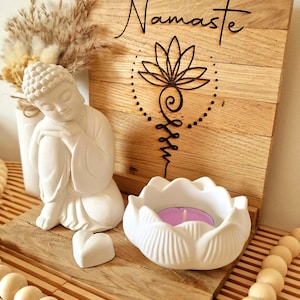 May include: A wooden plaque with the word Namaste and a black lotus symbol, featuring a white Buddha figurine, a white lotus-shaped candle holder with a purple tea light, a small white heart, and a dried floral arrangement on a wooden base.
